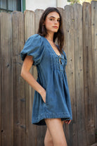 Woman wearing DELANEY soft denim puff sleeve denim romper featuring soft denim fabric, comfortable structured feel, deep V neckline, adjustable front tie detail, statement puff sleeves with gathered volume, pleated bodice, relaxed fit silhouette, and hidden side zipper closure.