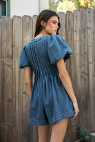 Woman wearing DELANEY soft denim puff sleeve denim romper featuring soft denim fabric, comfortable structured feel, deep V neckline, adjustable front tie detail, statement puff sleeves with gathered volume, pleated bodice, relaxed fit silhouette, and hidden side zipper closure.