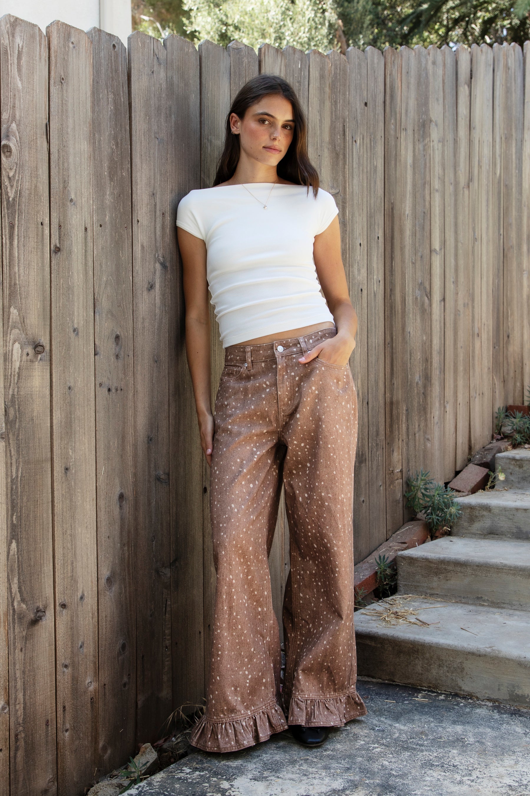 Woman wearing DOE deer printed ruffle hem wide leg denim pants featuring structured cotton print denim, high rise waist, button and zip fly closure, allover fawn inspired speckled print, wide leg silhouette, ruffle hem detail, classic five pocket design, and belt loops for western statement styling.