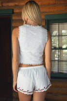 Women wearing TIC TOC JAIMY Poplin Embroidered Low Rise Shorts in whte with adjustable tie waistband and scalloped hem with floral embroidery paired with a white scalloped lace tie front tank top with brown boots in a cabin setting.