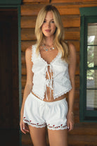Women wearing TIC TOC JAIMY Poplin Embroidered Low Rise Shorts in whte with adjustable tie waistband and scalloped hem with floral embroidery paired with a white scalloped lace tie front tank top with brown boots in a cabin setting.