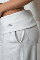 Woman wearing TIC TOC PJ31587BA COVE heather white wide leg lounge pants featuring a foldover waistband with logo detail, relaxed loose silhouette, wide leg shape, and side pockets, styled indoors with matching crop top and fur-trim hoodie.