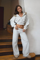 Woman wearing TIC TOC PJ31587BA COVE heather white wide leg lounge pants featuring a foldover waistband with logo detail, relaxed loose silhouette, wide leg shape, and side pockets, styled indoors with matching crop top and fur-trim hoodie.