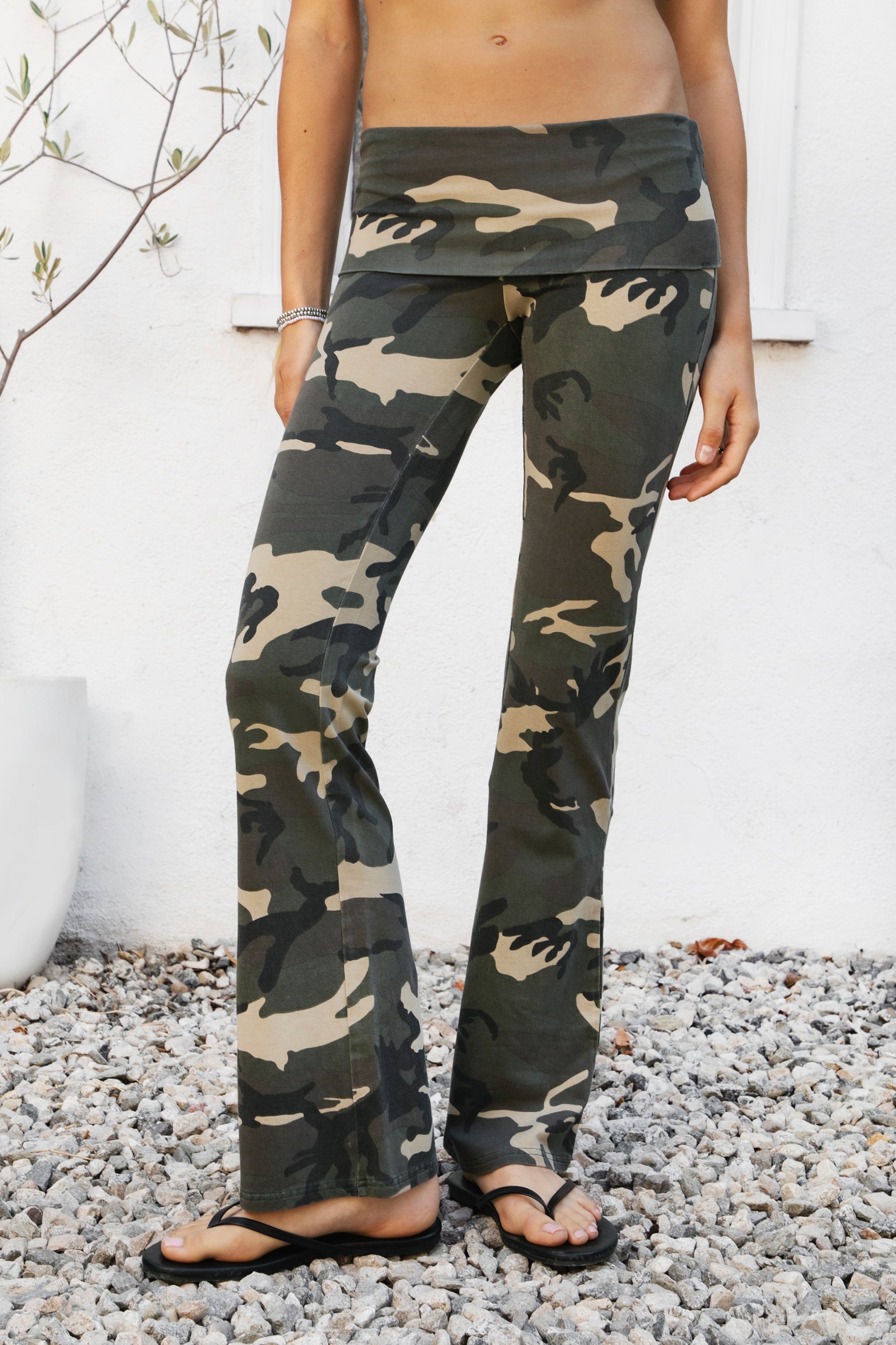 Woman wearing REMI dark military camo fold over flare leggings featuring soft stretch cotton jersey fabric, allover classic military camouflage print, low rise fit, wide fold over waistband, fitted hips and thighs, flared leg, garment washed finish, and full length pull on silhouette.