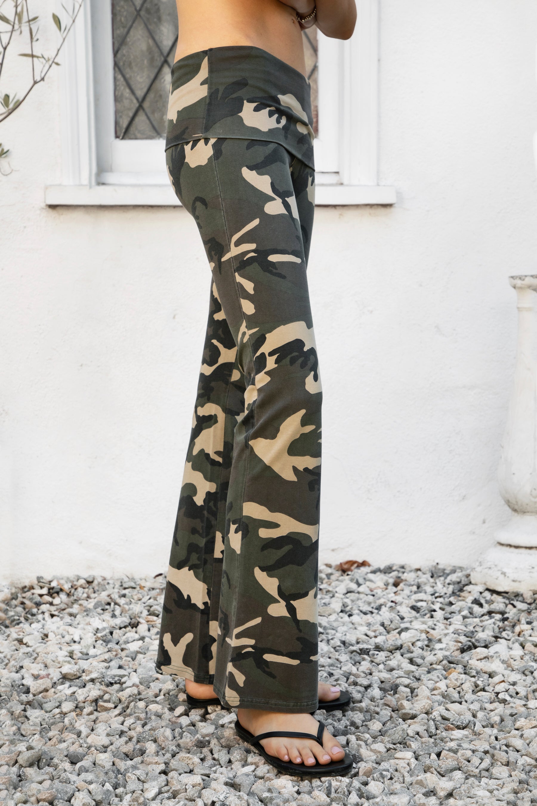 Woman wearing REMI dark military camo fold over flare leggings featuring soft stretch cotton jersey fabric, allover classic military camouflage print, low rise fit, wide fold over waistband, fitted hips and thighs, flared leg, garment washed finish, and full length pull on silhouette.