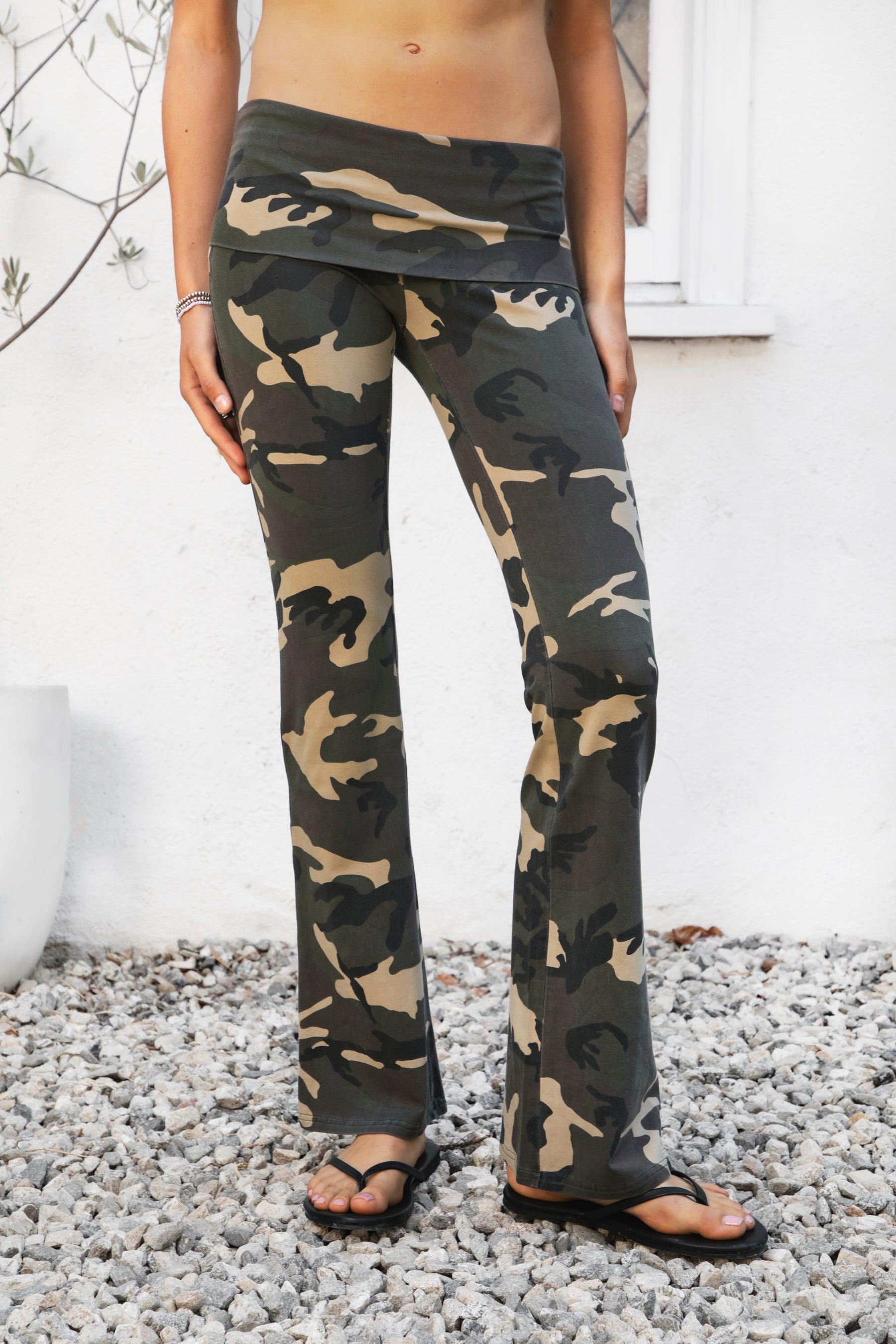 Woman wearing REMI dark military camo fold over flare leggings featuring soft stretch cotton jersey fabric, allover classic military camouflage print, low rise fit, wide fold over waistband, fitted hips and thighs, flared leg, garment washed finish, and full length pull on silhouette.