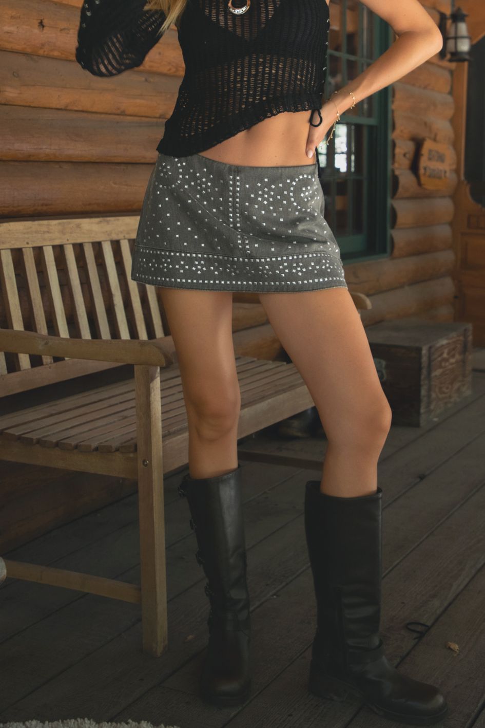 Person wearing a black top and studded gray skirt with knee-high boots on a wooden deck.