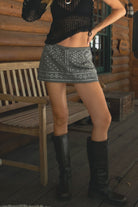 Person wearing a black top and studded gray skirt with knee-high boots on a wooden deck.