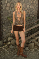 Woman wearing TIC TOC CARLEY Washed Denim Halter Top in brown with plunging neckline, lace-up torso, open back, and floral emboss detailing paired with bown suede shorts and cow boy boots agaist a stone wall. 