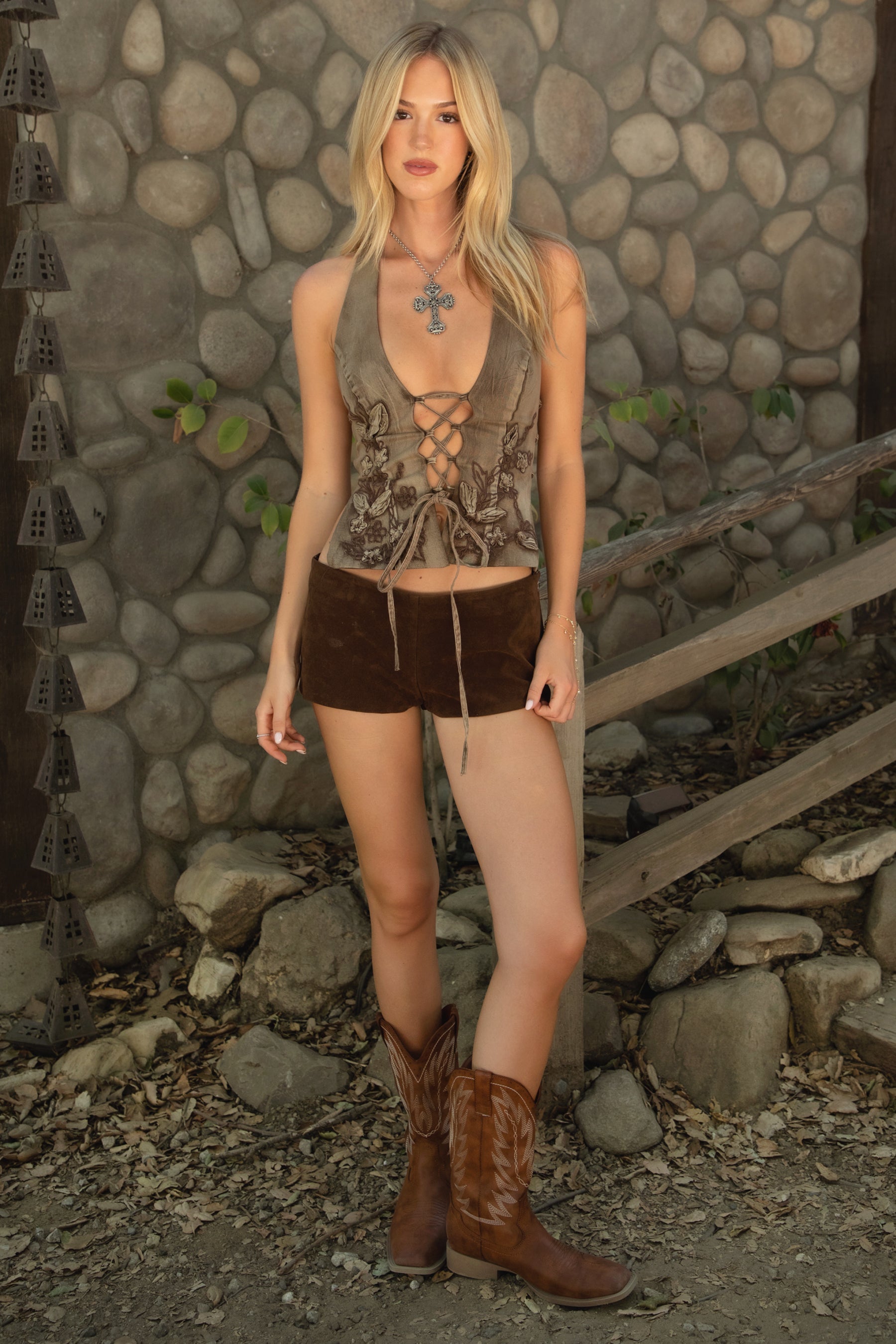 Woman wearing TIC TOC CARLEY Washed Denim Halter Top in brown with plunging neckline, lace-up torso, open back, and floral emboss detailing paired with bown suede shorts and cow boy boots agaist a stone wall. 