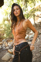 Woman wearing TIC TOC SIENNA Faux Suede Halter Bustier Top in tan with exposed eyelet lace detail and deep V neckline for a western festival look paired with denim pants on a forest setting. 