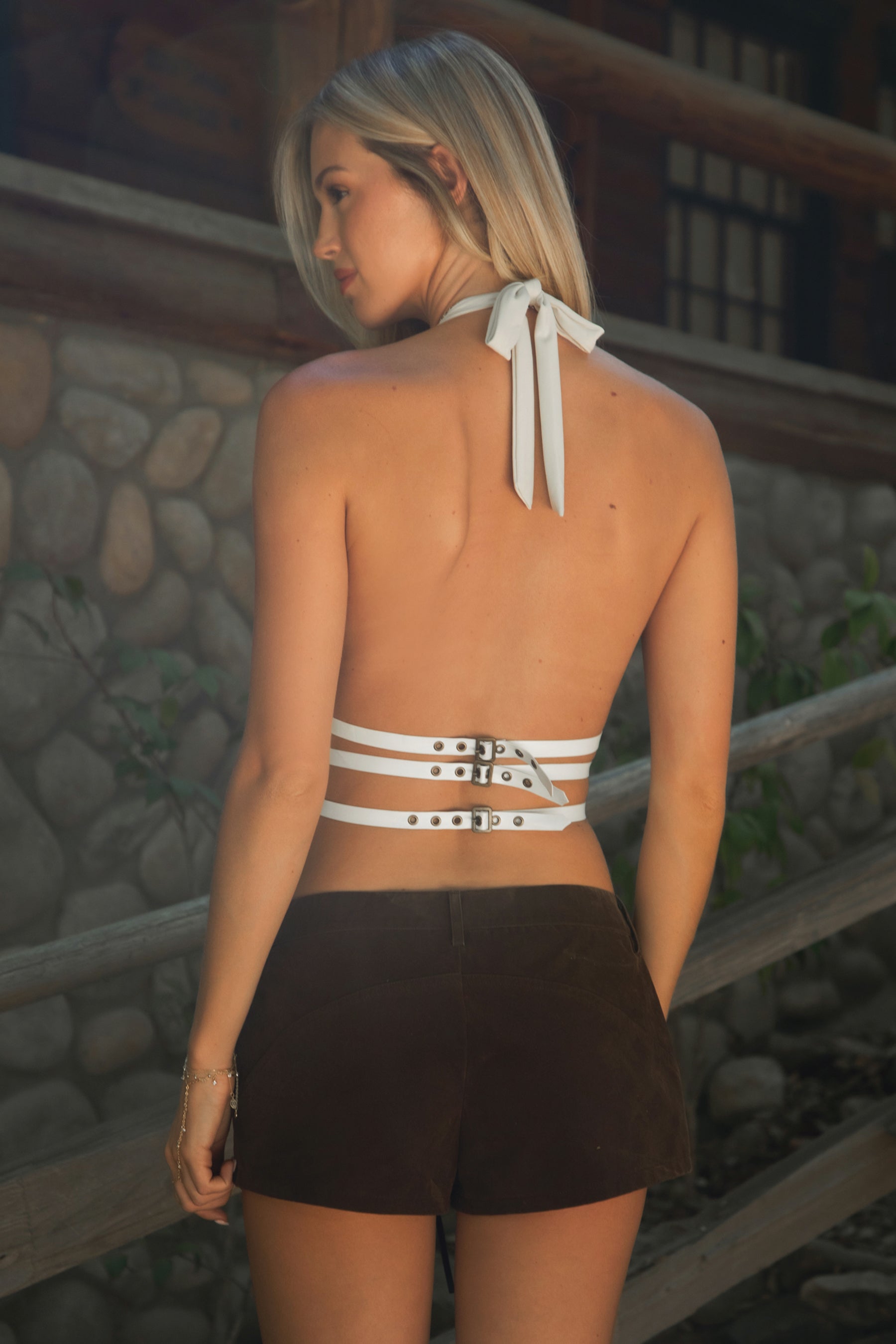 Woman wearing TIC TOC INDIE Split Front Halter Top in cream with plunging neckline, studded detailing, and open back with buckle straps and brown suede shorts outdoors. 