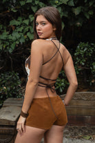 Woman wearing TIC TOC TA31807 MOOEDY-T BROWN halter fur corset top with a faux cow-pattern fur texture, plunging lace-up corset front, sculpted waist silhouette, and crisscross wraparound back ties styled with brown suede micro shorts in an outdoor rustic garden setting.
