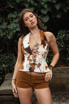 Woman wearing TIC TOC TA31807 MOOEDY-T BROWN halter fur corset top with a faux cow-pattern fur texture, plunging lace-up corset front, sculpted waist silhouette, and crisscross wraparound back ties styled with brown suede micro shorts in an outdoor rustic garden setting.