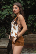 Woman wearing TIC TOC TA31807 MOOEDY-T BROWN halter fur corset top with a faux cow-pattern fur texture, plunging lace-up corset front, sculpted waist silhouette, and crisscross wraparound back ties styled with brown suede micro shorts in an outdoor rustic garden setting.