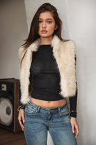 Woman wearing TIC TOC TA31834 BRIENNE faux fur vest in a natural tone; cropped open-front silhouette with plush texture, styled over a long-sleeve top and denim indoors.