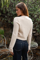 Woman wearing TIC TOC TA32002 RIEV oatmeal cropped knit cardigan with a soft brushed texture, delicate ribbon tie neckline, front button closure, and long ribbed-cuff sleeves paired with dark-wash denim jeans in a cozy garden setting.