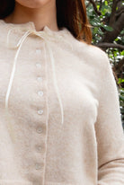 Woman wearing TIC TOC TA32002 RIEV oatmeal cropped knit cardigan with a soft brushed texture, delicate ribbon tie neckline, front button closure, and long ribbed-cuff sleeves paired with dark-wash denim jeans in a cozy garden setting.