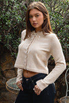 Woman wearing TIC TOC TA32002 RIEV oatmeal cropped knit cardigan with a soft brushed texture, delicate ribbon tie neckline, front button closure, and long ribbed-cuff sleeves paired with dark-wash denim jeans in a cozy garden setting.