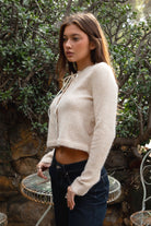 Woman wearing TIC TOC TA32002 RIEV oatmeal cropped knit cardigan with a soft brushed texture, delicate ribbon tie neckline, front button closure, and long ribbed-cuff sleeves paired with dark-wash denim jeans in a cozy garden setting.