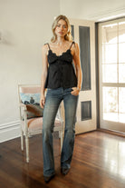 Woman wearing SOIE split V-neck satin cami top featuring scalloped lace trim along the neckline and front panel, adjustable self tie shoulder straps, five button front closure, front split hem detail, and an elastic back band for a soft feminine silhouette.