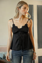 Woman wearing SOIE split V-neck satin cami top featuring scalloped lace trim along the neckline and front panel, adjustable self tie shoulder straps, five button front closure, front split hem detail, and an elastic back band for a soft feminine silhouette.