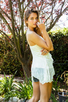Woman wearing RAVENA slub jersey lace V-neck ruffle cami top featuring lightweight slub jersey fabric, asymmetrical lace ruffle trim, adjustable bow tie neckline, handkerchief hem silhouette, adjustable straps, and soft jersey lining for feminine boho styling.