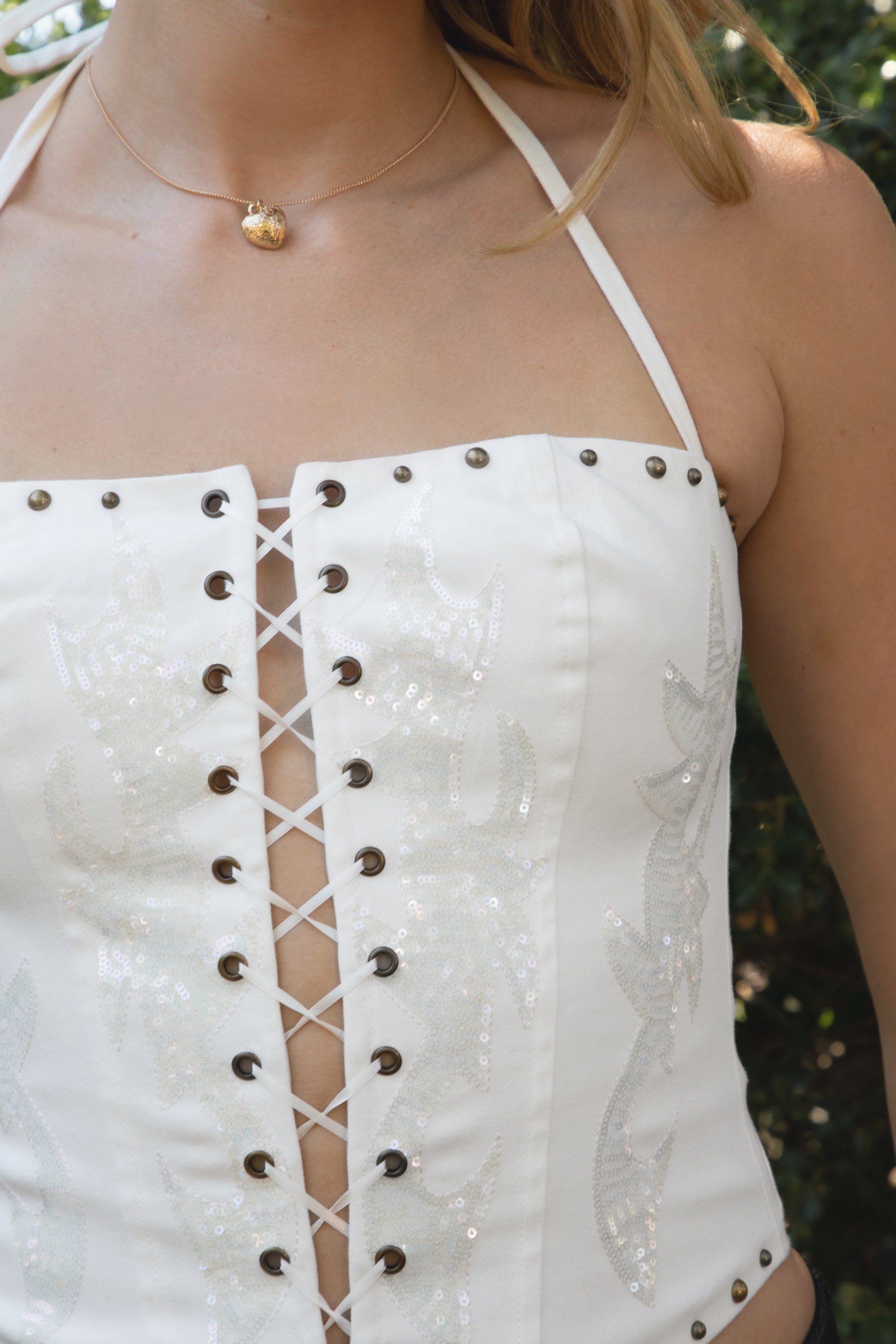 Woman wearing STARLANE sequin lace up halter denim top featuring a clear sequin embroidered front panel, eyelet lace up closure, thin adjustable halter straps, studded trim along the top and hem, and a fitted stretch denim silhouette with back zipper closure and jersey lining.