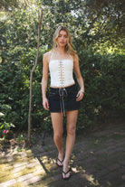 Woman wearing STARLANE sequin lace up halter denim top featuring a clear sequin embroidered front panel, eyelet lace up closure, thin adjustable halter straps, studded trim along the top and hem, and a fitted stretch denim silhouette with back zipper closure and jersey lining.