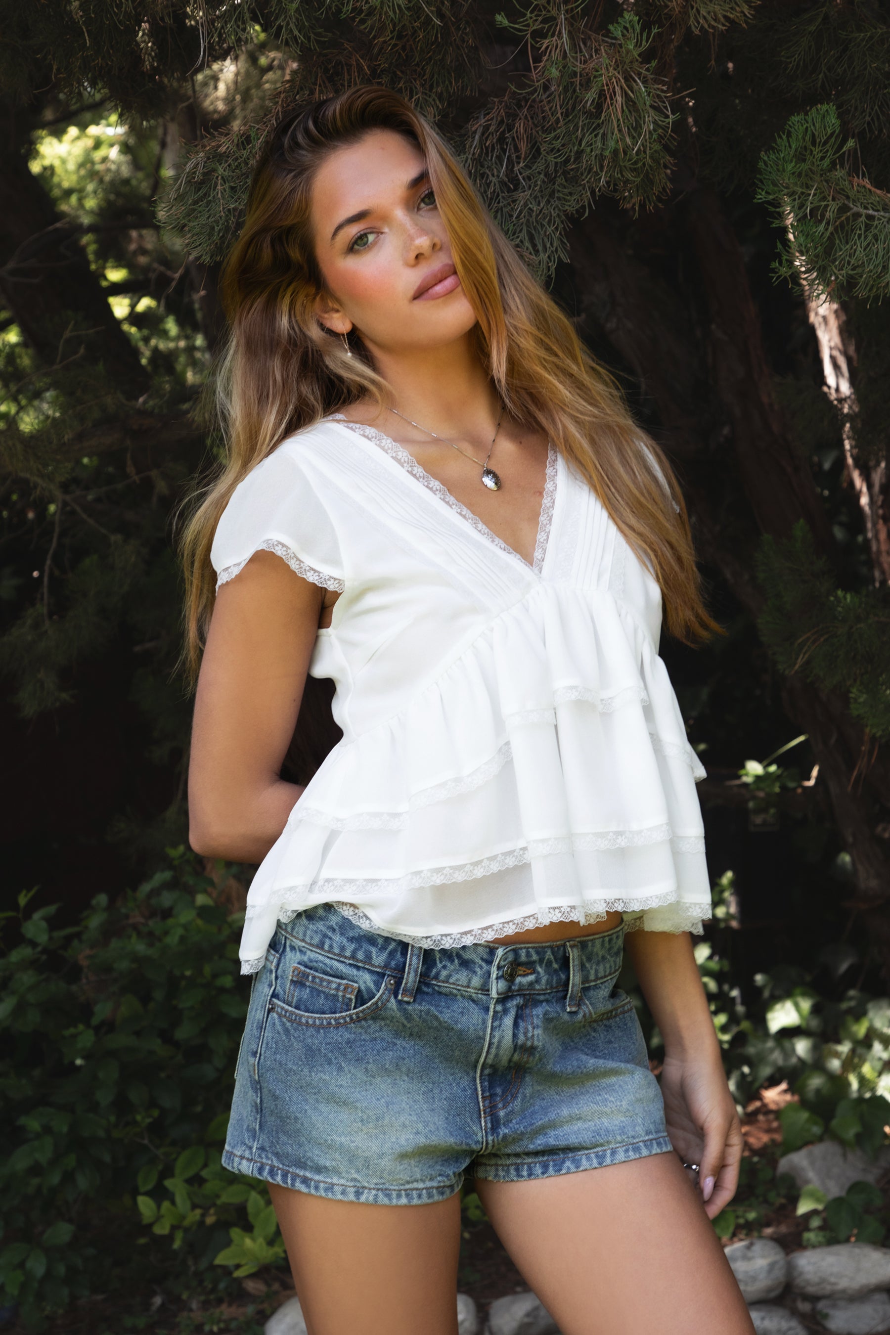 Woman wearing JULIETTE V-neck tiered ruffle short sleeve blouse featuring lightweight airy fabric, pleated V-neckline with lace trim, soft ruffle sleeves, tiered ruffle hem with lace detail, and a V-cut back with adjustable strap for a relaxed flowy silhouette.