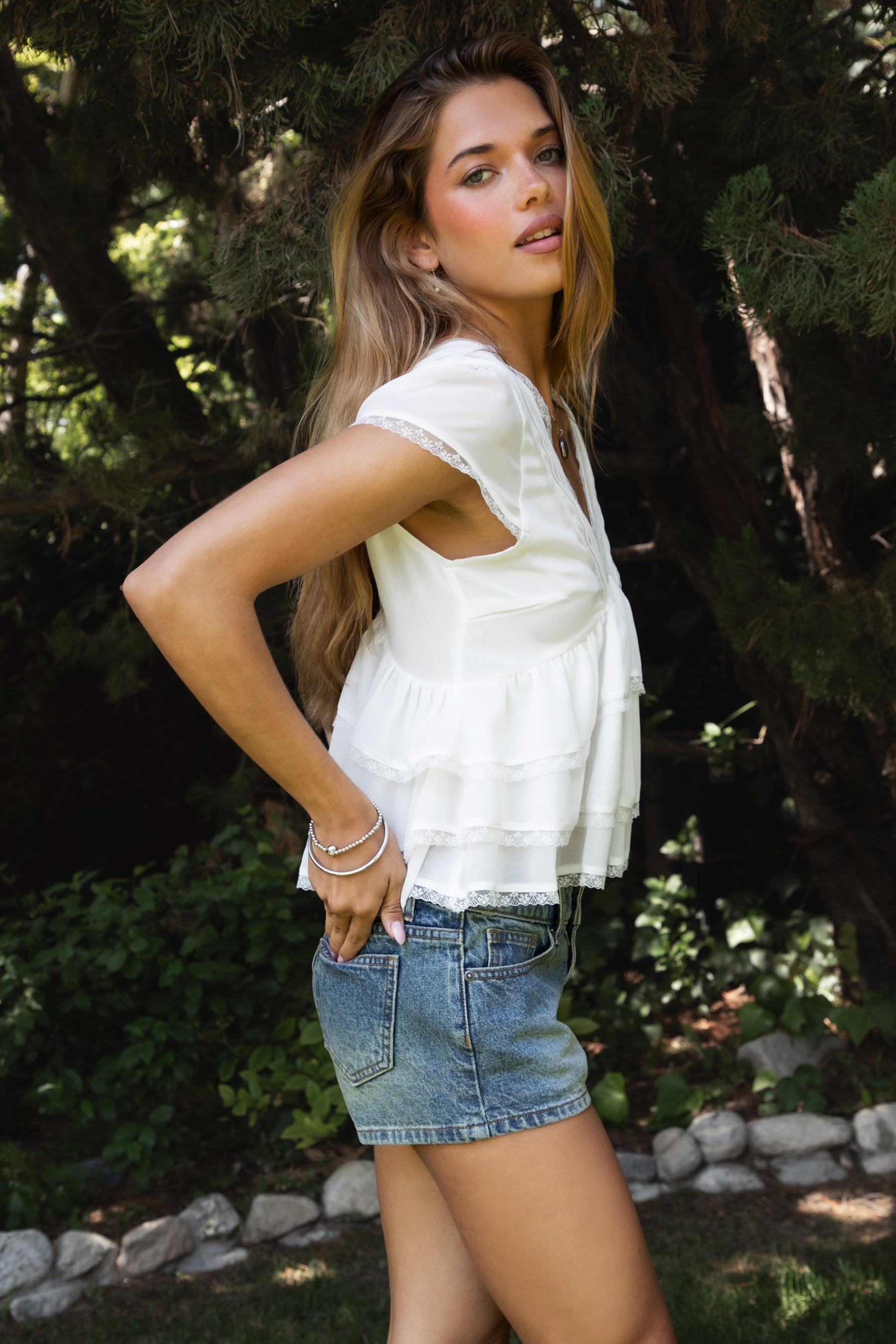 Woman wearing JULIETTE V-neck tiered ruffle short sleeve blouse featuring lightweight airy fabric, pleated V-neckline with lace trim, soft ruffle sleeves, tiered ruffle hem with lace detail, and a V-cut back with adjustable strap for a relaxed flowy silhouette.