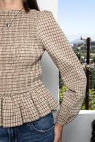 Woman wearing FARROW smocked micro plaid peplum blouse featuring lightweight cotton woven fabric, allover smocked bodice, classic boat neckline, feminine peplum hem, long puff sleeves with elastic cuffs, and soft neutral micro plaid pattern for timeless everyday styling.