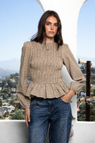 Woman wearing FARROW smocked micro plaid peplum blouse featuring lightweight cotton woven fabric, allover smocked bodice, classic boat neckline, feminine peplum hem, long puff sleeves with elastic cuffs, and soft neutral micro plaid pattern for timeless everyday styling.
