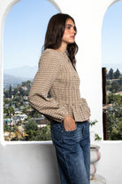 Woman wearing FARROW smocked micro plaid peplum blouse featuring lightweight cotton woven fabric, allover smocked bodice, classic boat neckline, feminine peplum hem, long puff sleeves with elastic cuffs, and soft neutral micro plaid pattern for timeless everyday styling.