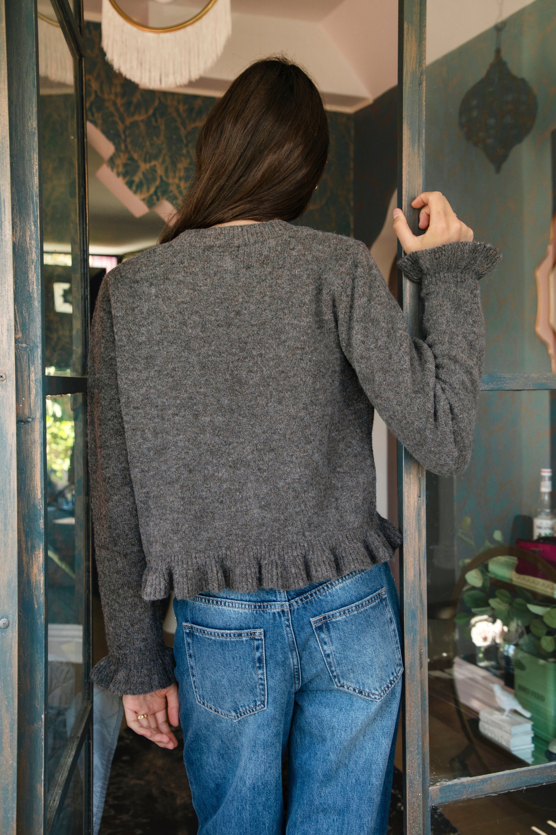 Woman wearing FRILLIA ruffle trim button front knit cardigan featuring soft brushed sweater knit fabric, cozy stretch feel, round neckline, button front closure, delicate ruffle trim along the front placket and hem, long sleeves with flared ruffle cuffs, and relaxed cropped silhouette.