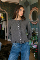 Woman wearing FRILLIA ruffle trim button front knit cardigan featuring soft brushed sweater knit fabric, cozy stretch feel, round neckline, button front closure, delicate ruffle trim along the front placket and hem, long sleeves with flared ruffle cuffs, and relaxed cropped silhouette.