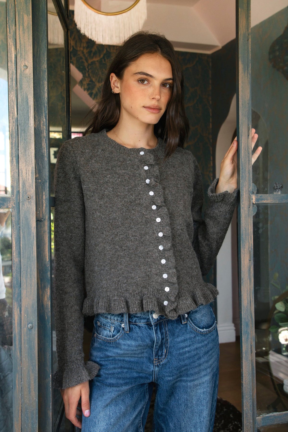 Woman wearing FRILLIA ruffle trim button front knit cardigan featuring soft brushed sweater knit fabric, cozy stretch feel, round neckline, button front closure, delicate ruffle trim along the front placket and hem, long sleeves with flared ruffle cuffs, and relaxed cropped silhouette.
