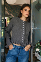 Woman wearing FRILLIA ruffle trim button front knit cardigan featuring soft brushed sweater knit fabric, cozy stretch feel, round neckline, button front closure, delicate ruffle trim along the front placket and hem, long sleeves with flared ruffle cuffs, and relaxed cropped silhouette.