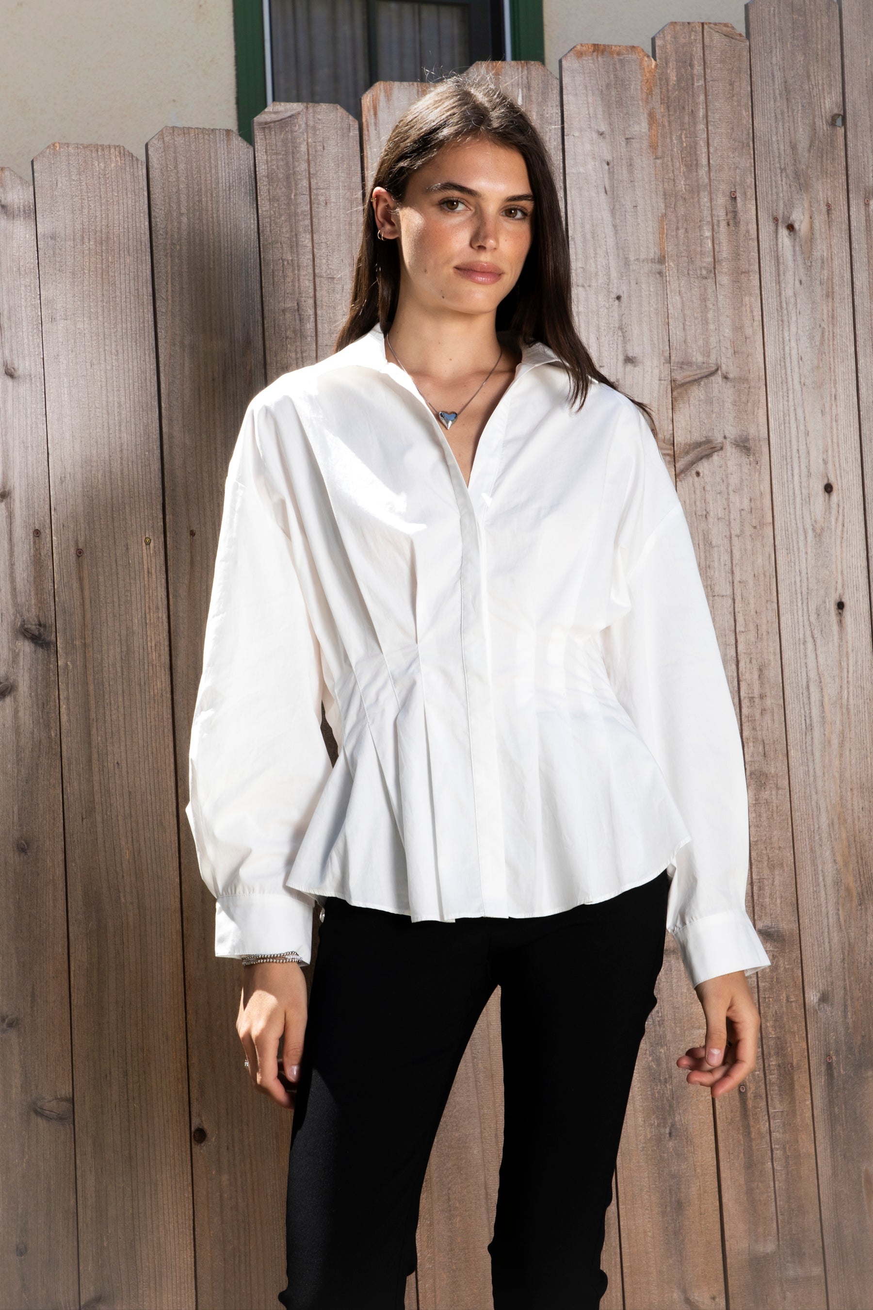 Woman wearing VIENNA tailored poplin peplum puff sleeve shirt featuring crisp cotton poplin fabric, structured feel, classic collared V neckline, hidden button front closure, defined waist seaming, soft pleated peplum hem, relaxed long balloon sleeves, and button cuffs for elevated everyday styling.