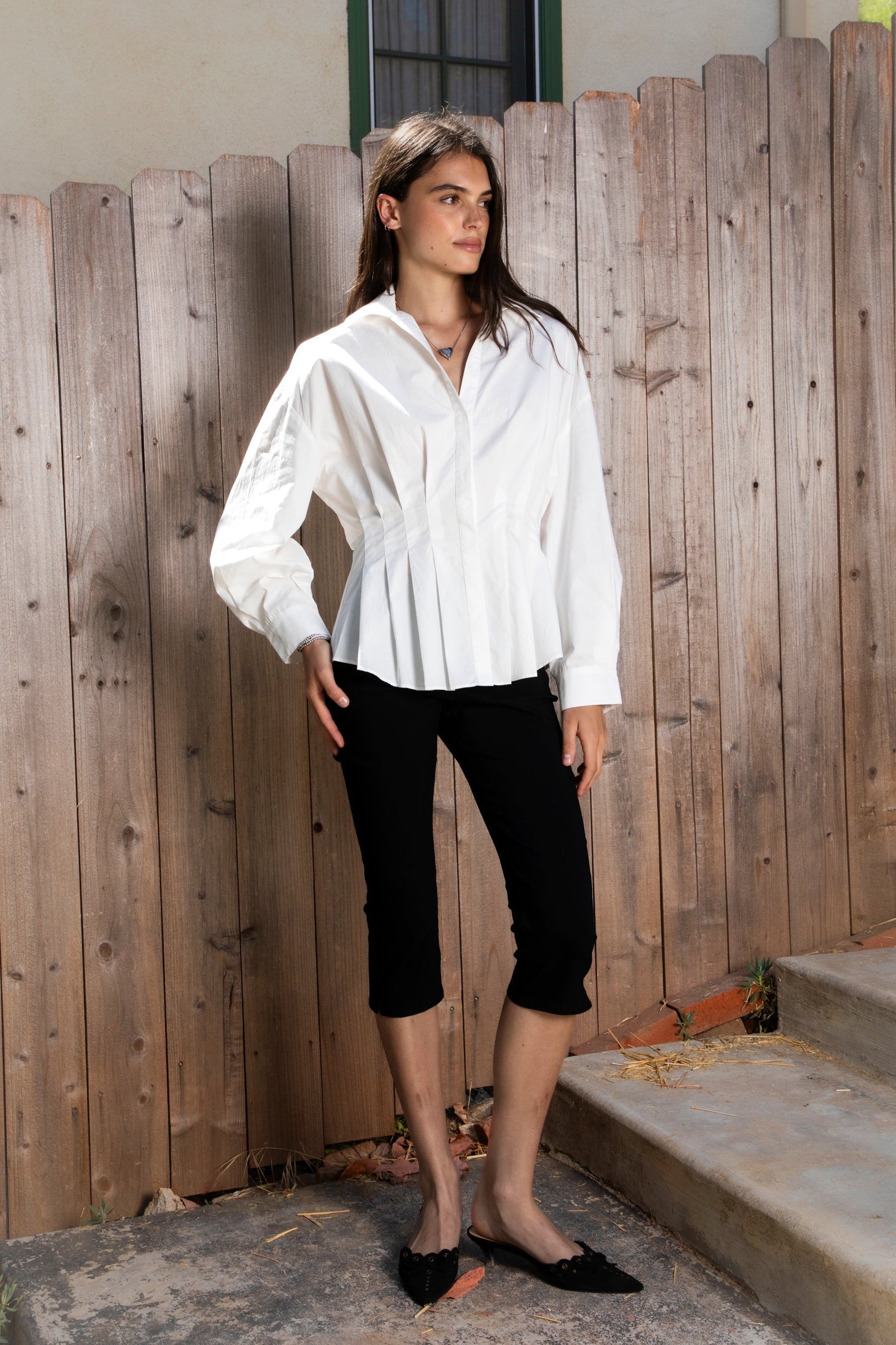 Woman wearing VIENNA tailored poplin peplum puff sleeve shirt featuring crisp cotton poplin fabric, structured feel, classic collared V neckline, hidden button front closure, defined waist seaming, soft pleated peplum hem, relaxed long balloon sleeves, and button cuffs for elevated everyday styling.