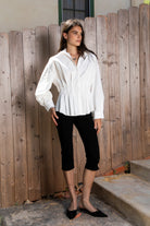 Woman wearing VIENNA tailored poplin peplum puff sleeve shirt featuring crisp cotton poplin fabric, structured feel, classic collared V neckline, hidden button front closure, defined waist seaming, soft pleated peplum hem, relaxed long balloon sleeves, and button cuffs for elevated everyday styling.