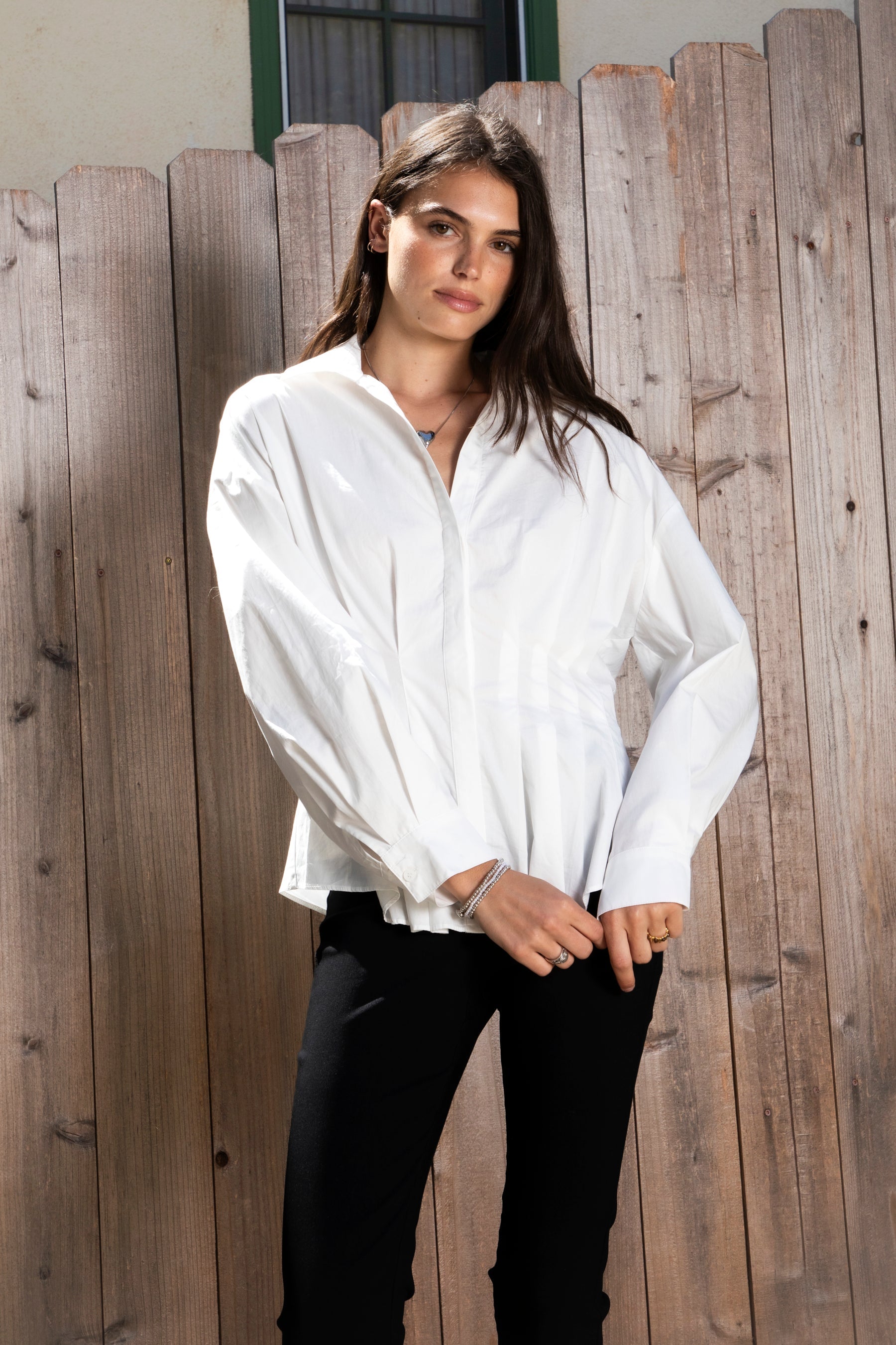 Woman wearing VIENNA tailored poplin peplum puff sleeve shirt featuring crisp cotton poplin fabric, structured feel, classic collared V neckline, hidden button front closure, defined waist seaming, soft pleated peplum hem, relaxed long balloon sleeves, and button cuffs for elevated everyday styling.