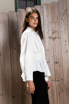Woman wearing VIENNA tailored poplin peplum puff sleeve shirt featuring crisp cotton poplin fabric, structured feel, classic collared V neckline, hidden button front closure, defined waist seaming, soft pleated peplum hem, relaxed long balloon sleeves, and button cuffs for elevated everyday styling.