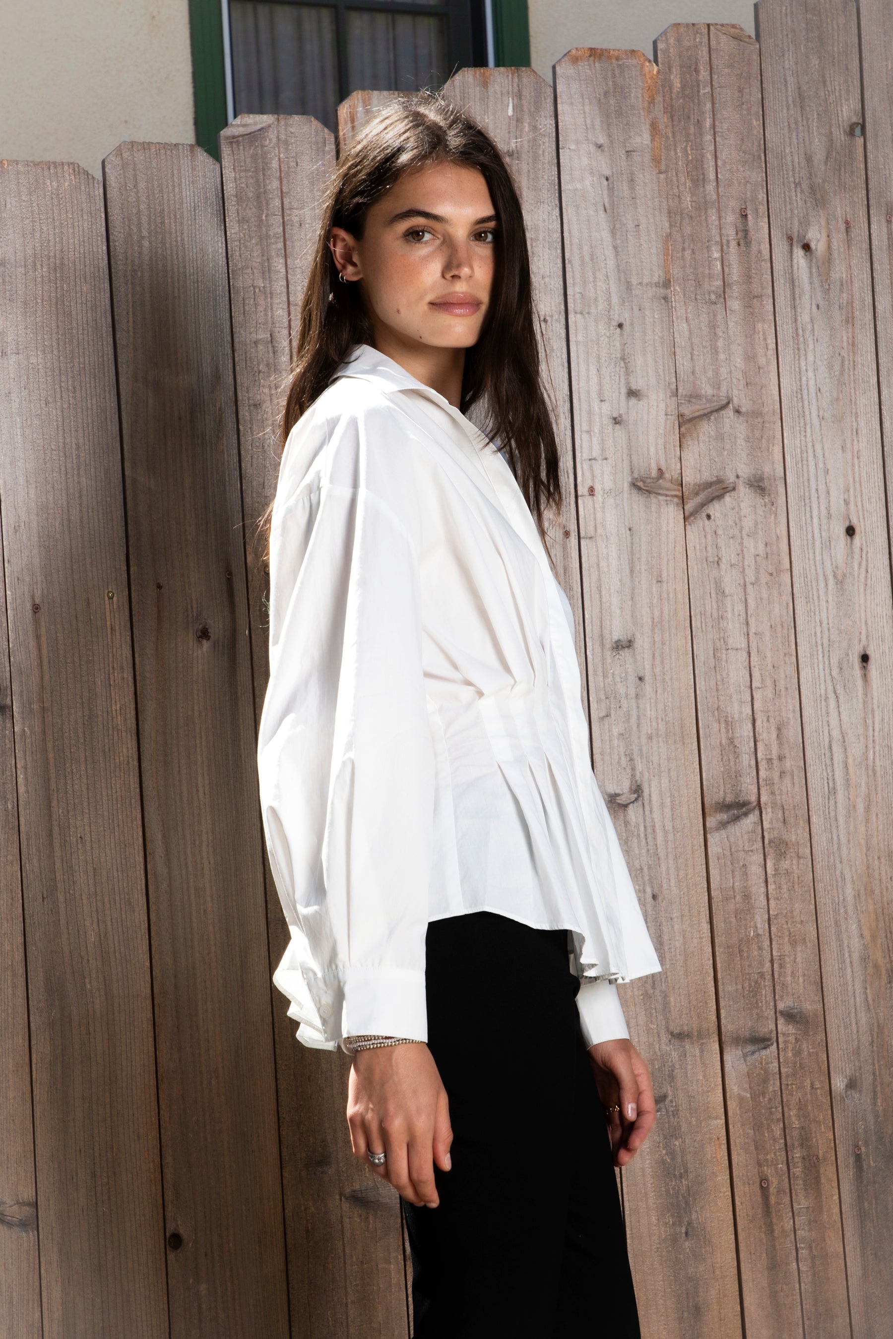 Woman wearing VIENNA tailored poplin peplum puff sleeve shirt featuring crisp cotton poplin fabric, structured feel, classic collared V neckline, hidden button front closure, defined waist seaming, soft pleated peplum hem, relaxed long balloon sleeves, and button cuffs for elevated everyday styling.