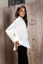 Woman wearing VIENNA tailored poplin peplum puff sleeve shirt featuring crisp cotton poplin fabric, structured feel, classic collared V neckline, hidden button front closure, defined waist seaming, soft pleated peplum hem, relaxed long balloon sleeves, and button cuffs for elevated everyday styling.