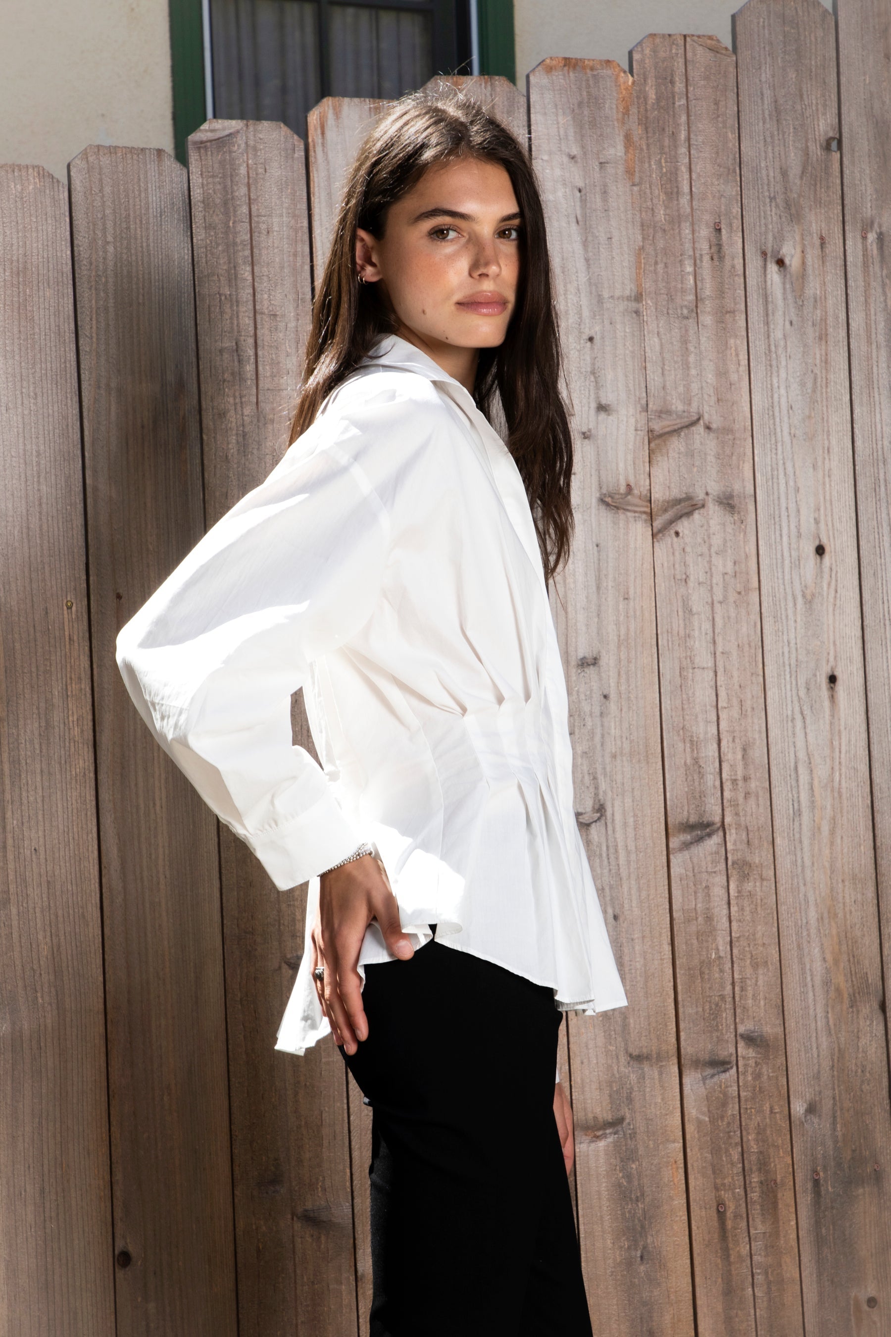 Woman wearing VIENNA tailored poplin peplum puff sleeve shirt featuring crisp cotton poplin fabric, structured feel, classic collared V neckline, hidden button front closure, defined waist seaming, soft pleated peplum hem, relaxed long balloon sleeves, and button cuffs for elevated everyday styling.