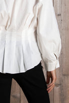 Woman wearing VIENNA tailored poplin peplum puff sleeve shirt featuring crisp cotton poplin fabric, structured feel, classic collared V neckline, hidden button front closure, defined waist seaming, soft pleated peplum hem, relaxed long balloon sleeves, and button cuffs for elevated everyday styling.
