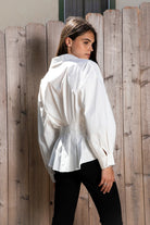 Woman wearing VIENNA tailored poplin peplum puff sleeve shirt featuring crisp cotton poplin fabric, structured feel, classic collared V neckline, hidden button front closure, defined waist seaming, soft pleated peplum hem, relaxed long balloon sleeves, and button cuffs for elevated everyday styling.
