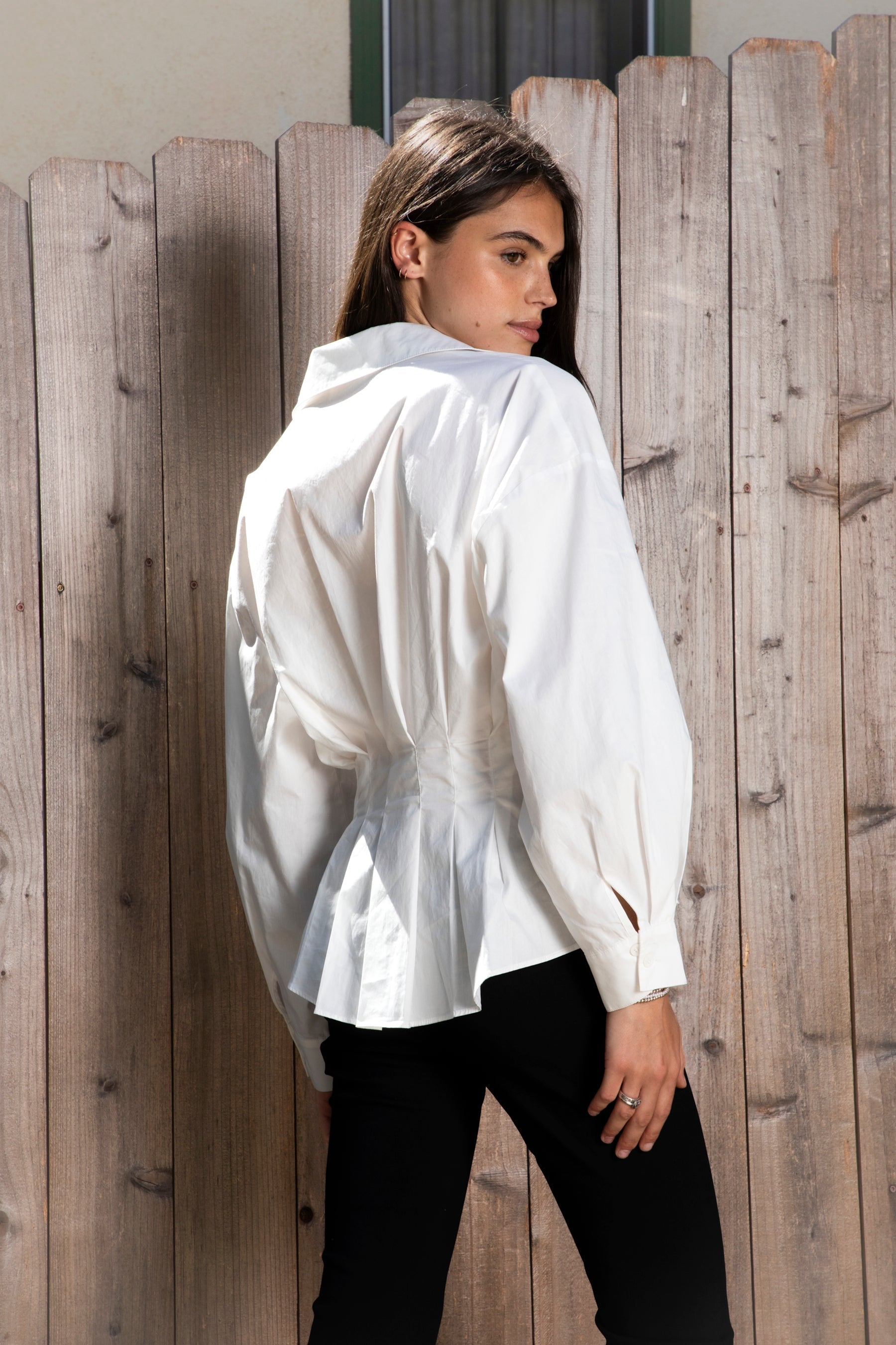 Woman wearing VIENNA tailored poplin peplum puff sleeve shirt featuring crisp cotton poplin fabric, structured feel, classic collared V neckline, hidden button front closure, defined waist seaming, soft pleated peplum hem, relaxed long balloon sleeves, and button cuffs for elevated everyday styling.