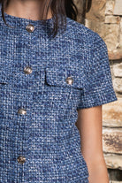 Woman wearing COCO navy blue lurex tweed short sleeve top featuring shimmering textured tweed fabric, subtle metallic lurex detail, round neckline, fitted structured bodice, cropped length, elegant gold point buttons, decorative faux chest pockets, and fully lined construction designed to coordinate with the matching COCO skort.